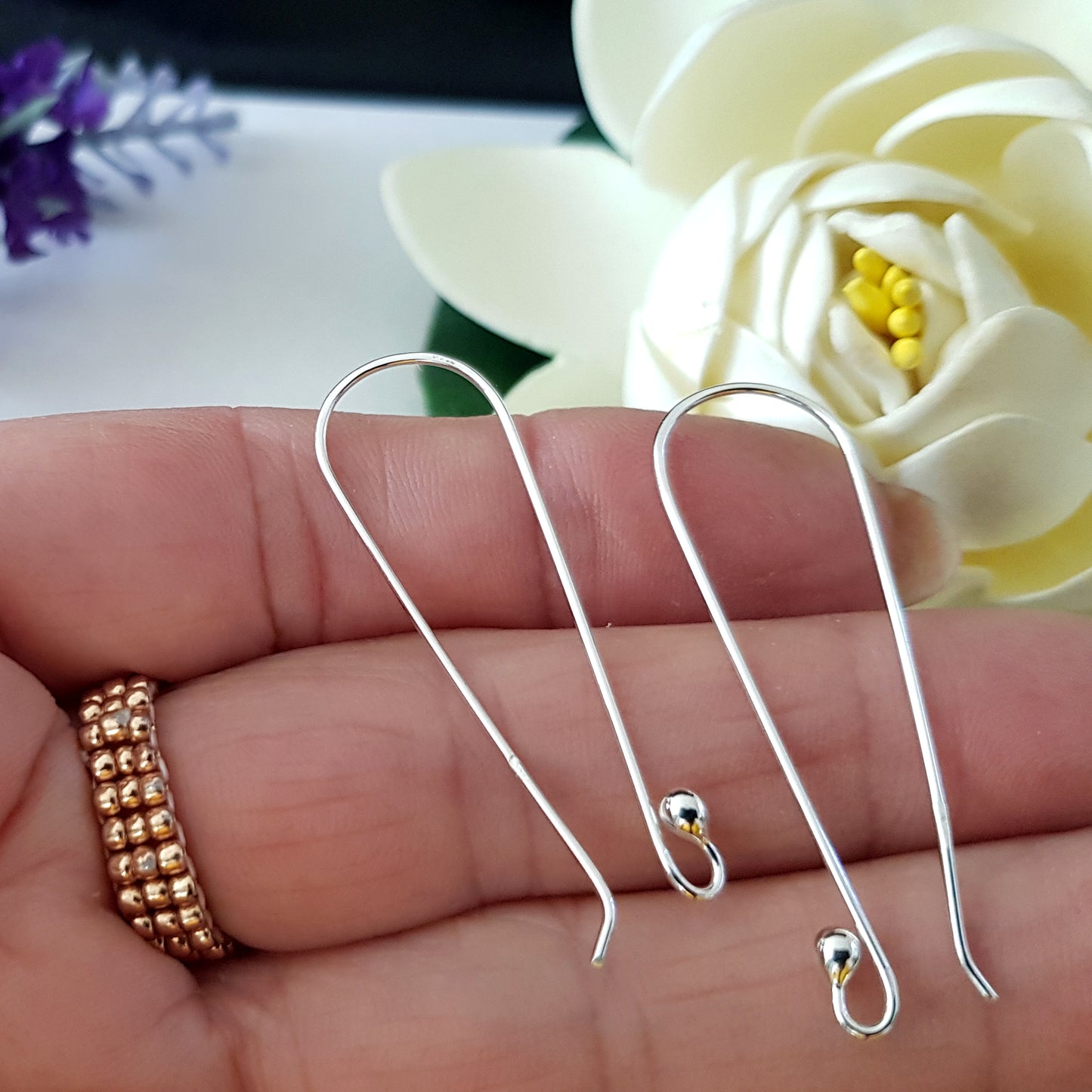 A pair of handmade sterling silver ear hooks displayed on a person's hand, with a flower in the background.