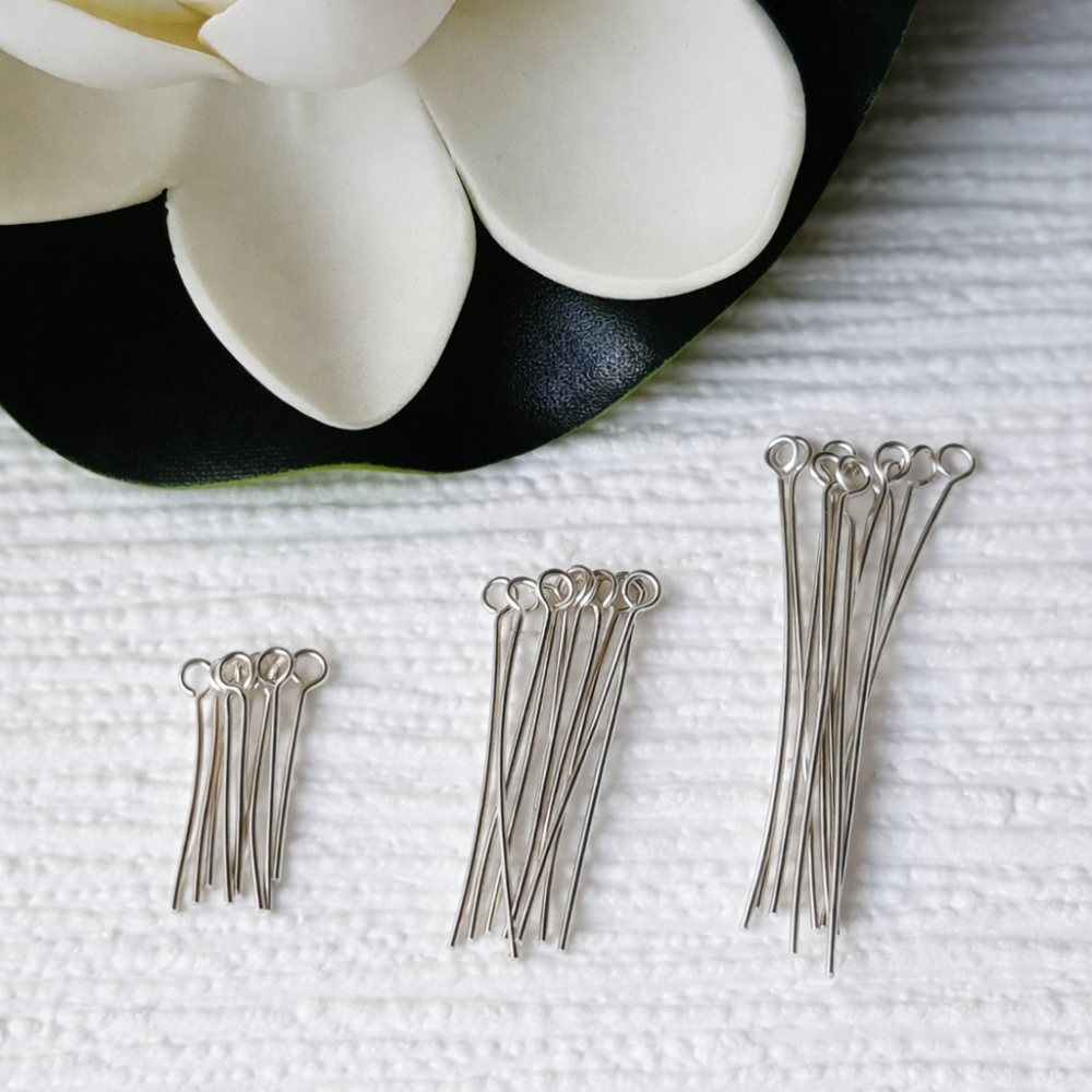 Comparison lengths, Silver eye pins on a textured white surface with a black plate and white flowers in the background.