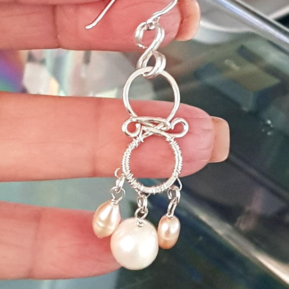 Close-up of handmade chandelier pearl earring held in hand, showing intricate sterling silver wirework with white and soft-pink freshwater pearls – FKJ-210E by Kalitheo