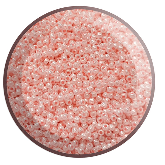 A round container filled with baby pink opaque luster Toho seed beads. circle white background